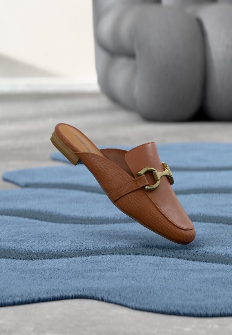Mocassin en cuir marron à enfiler avec détail mors doré, posé sur un tapis bleu texturé avec un meuble gris coussiné en arrière-plan.