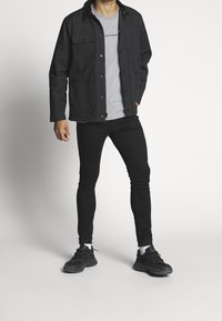 Hombre vistiendo una chaqueta negra, una camiseta gris de Calvin Klein, pantalones slim negros, calcetines blancos y zapatillas negras, de pie con una mano en el bolsillo.
