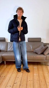 Zwarte pufferjas met een hoge kraag en rits, gecombineerd met lichtblauwe spijkerbroek en glanzende donkere schoenen. Staand op een houten vloer.