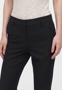 Femme portant un pantalon noir sur mesure et un haut sans manches côtelé beige clair, debout, les mains détendues le long des hanches.