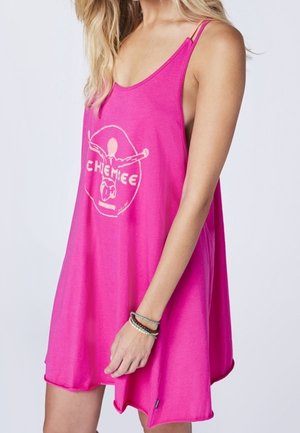 Žena v volných, jasně růžových šatech bez rukávů s tenkými ramínky a bílým grafickým logem na přední straně.