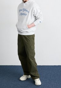 Persona vestida con sudadera clara de "New Balance Athletics", pantalones cargo verde olivo y zapatillas blancas, de pie sobre una alfombra azul.