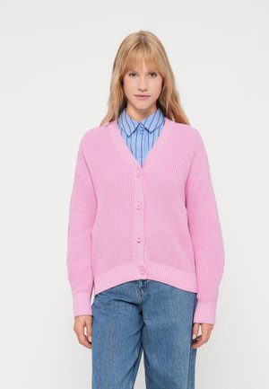 Cardigan tricoté rose avec un col en V, cinq boutons sur le devant et des poignets côtelés. Porté par-dessus une chemise à boutons bleu à rayures.