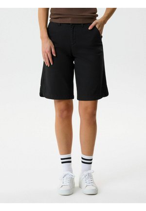 Person trägt schwarze knielange Shorts, weiße Turnschuhe und weiße Socken mit zwei schwarzen Streifen, steht mit Händen in und nahe den Taschen.