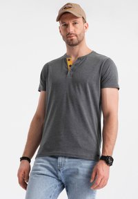 Camiseta henley gris de manga corta con una tapeta de botones amarilla, combinada con jeans azul claro. Una gorra color beige completa el atuendo.