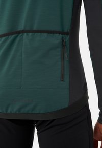 Veste de sport zippée verte et noire avec une texture finie, un matériau extensible et un logo au bas. Présente des panneaux de contraste.