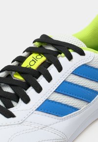 Chaussure de sport blanche avec des bandes bleues, des lacets noirs, et un détail sur la languette en jaune néon avec un texte noir, vue en gros plan.