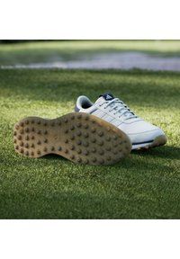 Chaussure de golf Adidas blanche sur l'herbe, montrant la semelle avec des crampons en caoutchouc et la tige avec des lacets et le logo sur la languette.