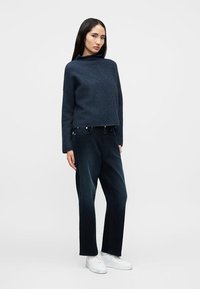 Γυναίκα που στέκεται φορώντας ένα σκουρομπλε πουλόβερ με ζιβάγκο, άνετο μαύρο τζιν και λευκά αθλητικά παπούτσια σε ένα απλό ανοιχτόχρωμο φόντο.