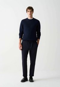 Námořnicky modrý pletený svetr s kulatým výstřihem, spojený s námořnicky modrými tepláky s elastickým pasem a šňůrkou. Lesklé černé boty.