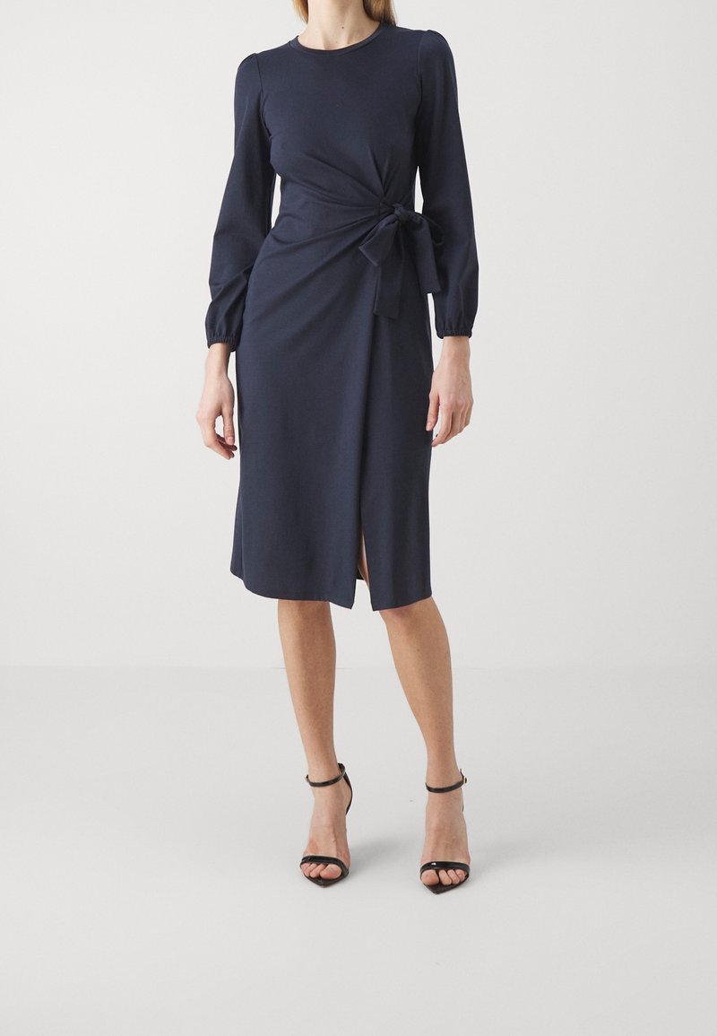 Robe portefeuille bleu marine à manches longues, avec une ceinture à la taille, une fente à l'avant et un tissu lisse et léger. Des talons complètent la tenue.