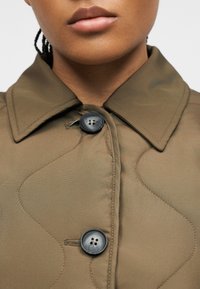 Κοντινή λήψη ενός ατόμου που φοράει ένα καφέ καπιτονέ μπουφάν με γιακά και δύο μεγάλα σκούρα κουμπιά δένονται μπροστά.