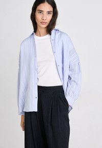 Camicia a righe azzurro chiaro con bottoni e vestibilità morbida, indossata sopra una t-shirt bianca. Abbinata a pantaloni scuri a righe sottili, che mostrano texture stratificate.