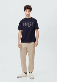 Mornarsko modra bombažna majica s tiskom rastlin, v kombinaciji s svetlo bež hlačami in belimi teniskami. Preprost dizajn s kratkimi rokavi.