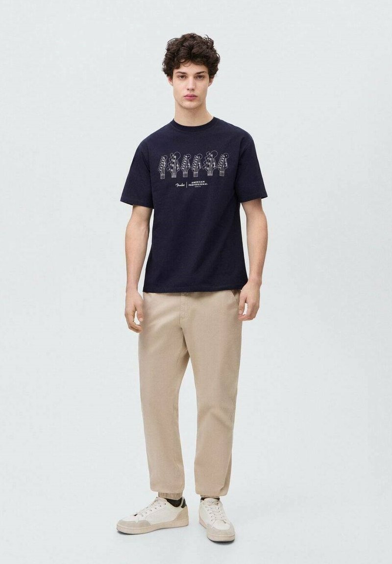 Mornarsko modra bombažna majica s tiskom rastlin, v kombinaciji s svetlo bež hlačami in belimi teniskami. Preprost dizajn s kratkimi rokavi.