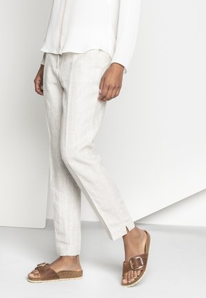 Person iført lyse beige linnedbukser, hvid bluse med lange ærmer og brune ruskindssandaler med stor spænde, stående på lyst gulv.