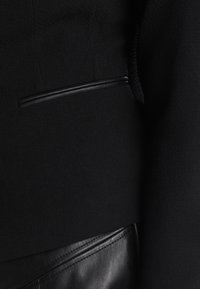 Veste noire avec un tissu texturé et un accent de poche en cuir lisse. Le design comprend une coupe élégante et ajustée ainsi que des détails subtils.