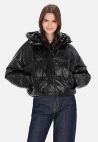 Fekete, fényes hatású pufidzseki cipzáras elülső résszel, levehető kapucnival és oldalzsebekkel. Fehér felső és kék farmer felett viselve.