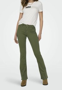 Jean évasé vert avec une texture lisse, associé à un t-shirt blanc arborant un design de lettres noires, et des bottines brunes à la cheville.