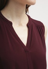 Burgundy sleeveless blouse with a V-neckline, featuring soft, lightweight fabric and subtle pleating detail near the shoulders.