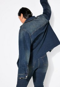 Chaqueta de denim en azul oscuro con un lavado en azul claro en los hombros, corte relajado y puños con botones, combinada con jeans de denim oscuros a juego.