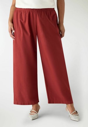 Persona che indossa pantaloni a vita alta e gamba larga color ruggine con fascia elastica in vita e scarpe slingback bianche con punte appuntite e borchiate.