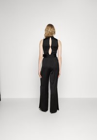 Mono de satén negro con un cuerpo ajustado, detalle de escote en la espalda, pantalones de pierna ancha y un cinturón a juego en la cintura. Textura suave.