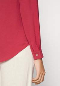 Camicia rossa a maniche lunghe con polsino abbottonato e orlo curvo, realizzata in un tessuto morbido e testurizzato. Indossata con pantaloni in lino di colore chiaro.