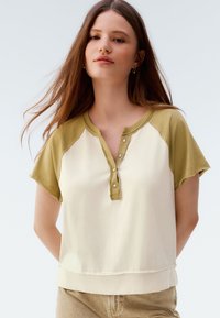 Short-sleeve top with a cream body and olive green raglan sleeves. Features a buttoned henley neck and a relaxed fit. Soft fabric texture.