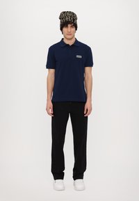 Mornarsko majico s kratkimi rokavi, ovratnikom in vezenim logotipom, ujemajočo se s črnimi hlačami in vzorčeno pleteno kapo. Bele superge.