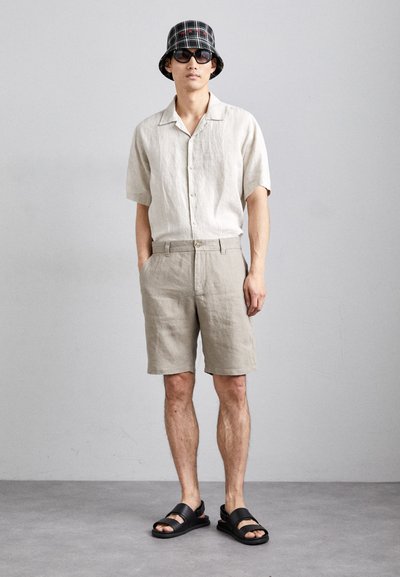 Chemise en lin à manches courtes beige clair, assortie à un short beige et des sandales à glissière noires. Accessoirisée avec un chapeau seau à carreaux.