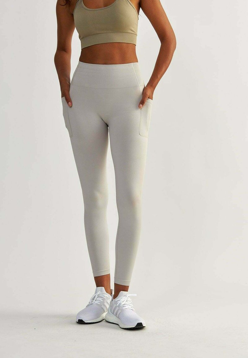 Leggings grigi chiaro con vita alta e tasche laterali. Realizzati in materiale elastico, abbinati a scarpe da ginnastica bianche e un top corto beige.