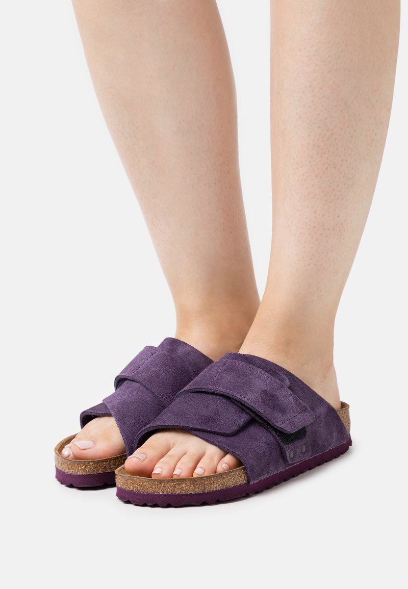 Füße in lila Wildleder-Slippern mit verstellbaren Riemen und Kork-Fußbetten auf weißem Hintergrund.