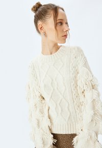 Biały sweter dzianinowy z wzorem w warkocze i frędzlami na rękawach. Posiada okrągły dekolt i ściągacz na dole. Modelka nosi spódnicę w brązowy wzór.