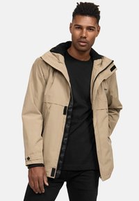 Beige waterdichte jas met een capuchon en zwarte accenten, voorzien van een voorrits, twee grote zakken en verstelbare manchetten.