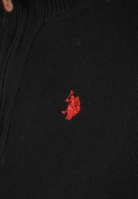 Red embroidered polo player logo on black knit fabric with visible zipper seam.
