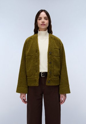 Vrouw met een olijfgroene getextureerde jas met knopen over een witte coltrui en donkerbruine broek met zwarte riem.