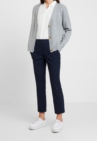 Světle šedý cardigan se zlatými knoflíky, bílá košile s knoflíky, tmavě modré kostkované kalhoty a bílé tenisky, což představuje neformální, vrstvený outfit.