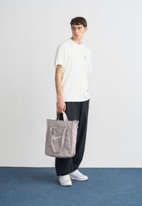 Γκρι τσάντα ώμου από νάιλον με ασημένια μεταλλικά στοιχεία, που φέρει μεγάλο λογότυπο Nike. Το μοντέλο φοράει λευκό μπλουζάκι και μαύρο παντελόνι, στέκεται πάνω σε μπλε χαλί.