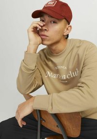 Beige sweatshirt with "Marina Club" print, ribbed cuffs, paired with a maroon cap featuring a logo patch. Model sits on a wooden chair.