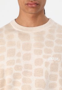 Beige gestrickter Pullover mit einem strukturierten Muster aus hellbraunen Formen und einem weißen gestickten Logo auf der linken Seite der Brust.