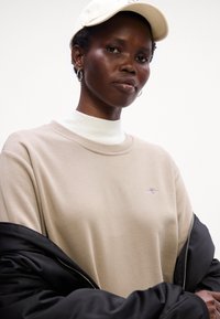 Beiger Sweatshirt mit Rundhalsausschnitt, strukturiertem Stoff, gesticktem Logo auf der Brust, kombiniert mit einer schwarzen Pufferjacke und einem weißen Rollkragenpullover darunter.