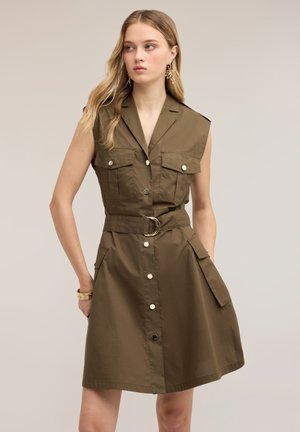 Jeune femme portant une robe sans manches vert olive ceinturée avec des boutons sur le devant et des poches à rabat, debout devant un fond neutre uni.