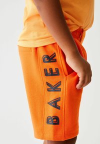 Orange bomuldsbukser med sidelomme og fed sort "BAKER" tekstdesign. Livremmen er elastisk for komfort og pasform.