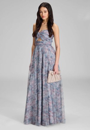 Kobieta ubrana w niebiesko-różową kwiecistą suknię maxi bez ramiączek z wycięciem, trzymająca beżową plisowaną kopertówkę, stojąca na jednolitym tle.