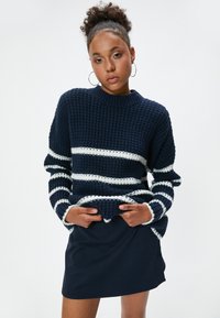 Navy gestrickter Pullover mit weißen horizontalen Streifen, geripptem Rundhalsausschnitt und strukturiertem Muster. Kombiniert mit einem dunkelblauen Minirock.