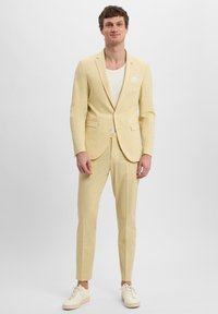 Helle gelbe Anzug aus strukturiertem Stoff, mit einem taillierten Blazer mit zwei Vorderaschen und passenden Hosen. Kombiniert mit weißen Sneakers.