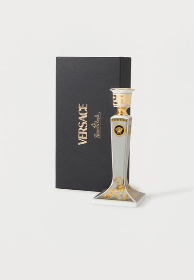 Valkoinen ja kullanvärinen koristeellinen Versacen kynttilänjalka Medusa-emblemalla, vieressä musta Versacen rasia kultaisilla kirjaimilla.