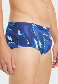 Niebieskie kąpielówki w wzory z granatowym tłem i abstrakcyjnymi niebieskimi oraz białymi kształtami, wykonane z gładkiego, elastycznego materiału.