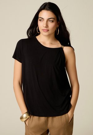 Woman with long dark hair wearing a black asymmetrical top with a gold clasp on one shoulder, gold hoop earrings, and brown pants, hands in pockets.
